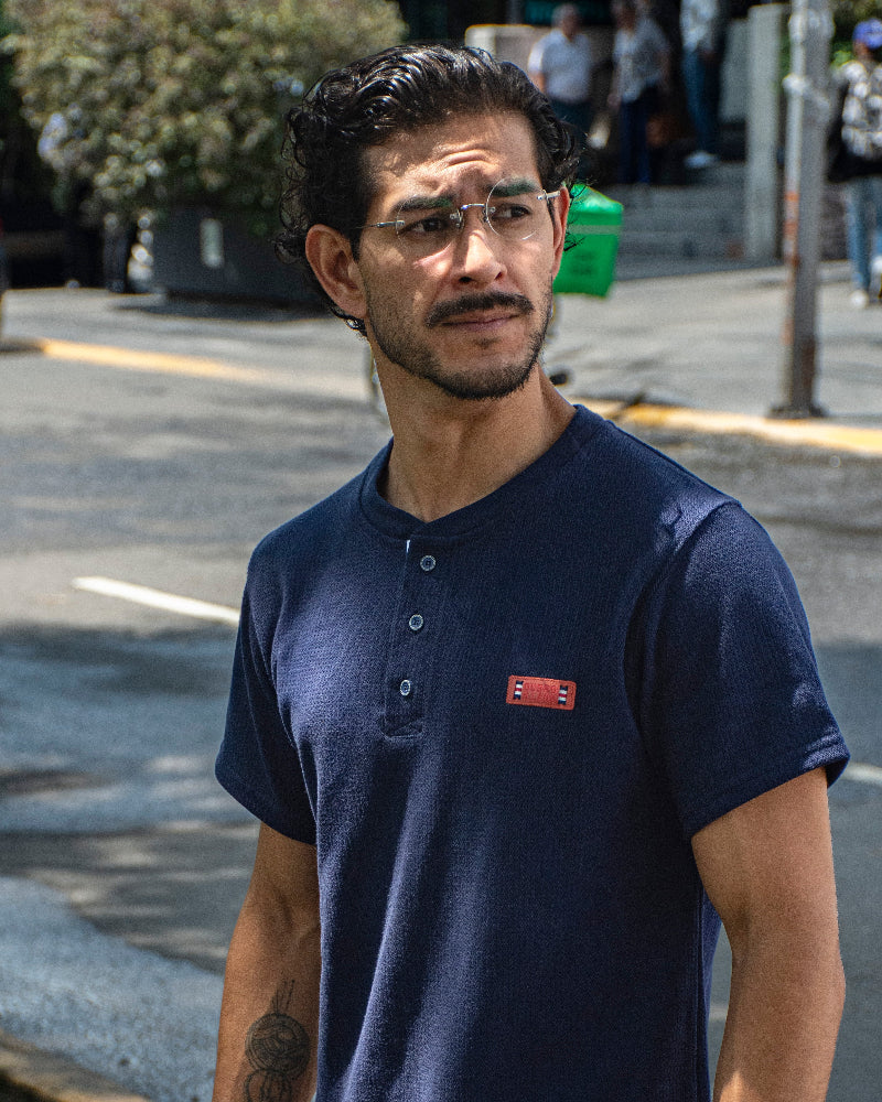 Hombre con gafas de sol y reloj de perfil en un parque, vistiendo una camiseta tipo Henley azul marino texturizada de la marca Dubrovnik con botones abiertos