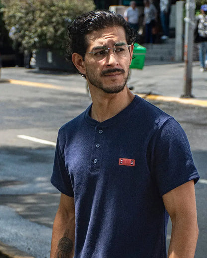 Hombre con gafas de sol y reloj de perfil en un parque, vistiendo una camiseta tipo Henley azul marino texturizada de la marca Dubrovnik con botones abiertos