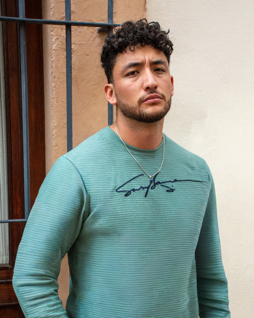 Plano medio de hombre con camiseta verde agua de textura acanalada y firma gráfica en el pecho, posando frente a una pared rústica