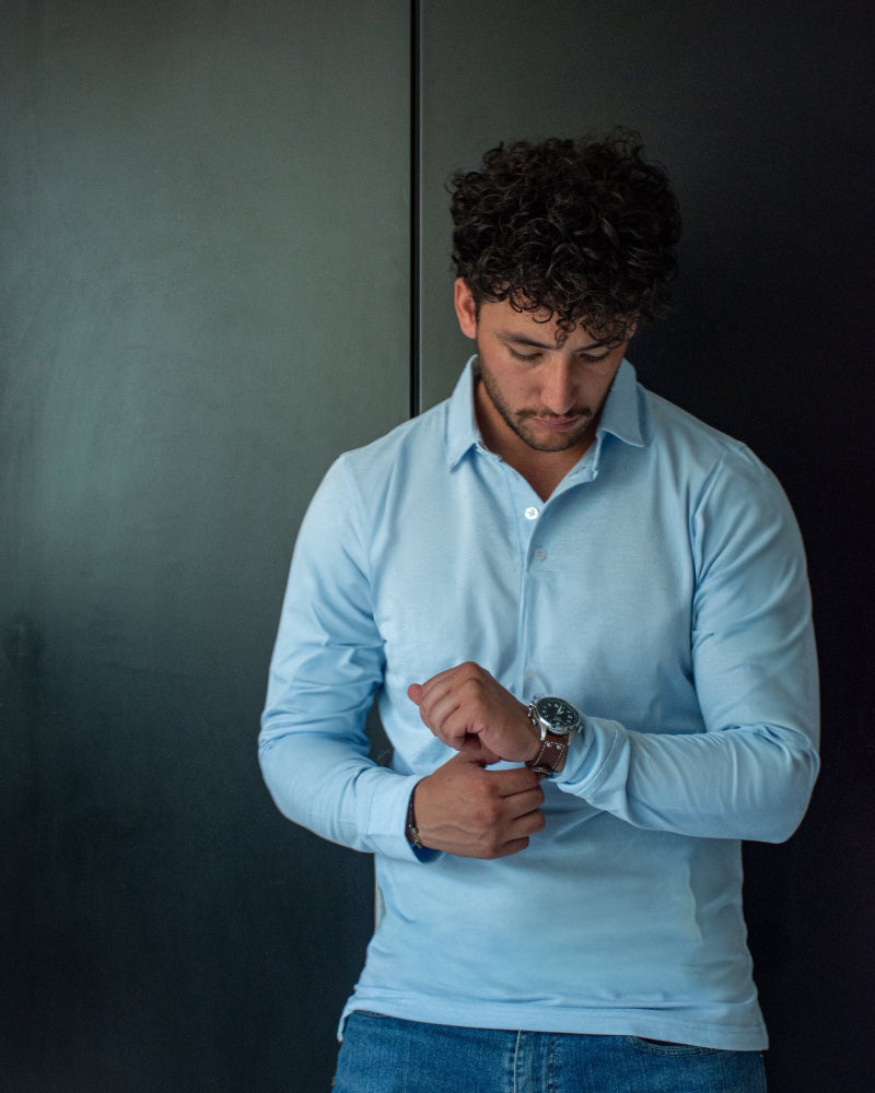 Hombre con polo de manga larga en color celeste claro, ajustándose un reloj de pulsera, con estilo casual y fondo oscuro