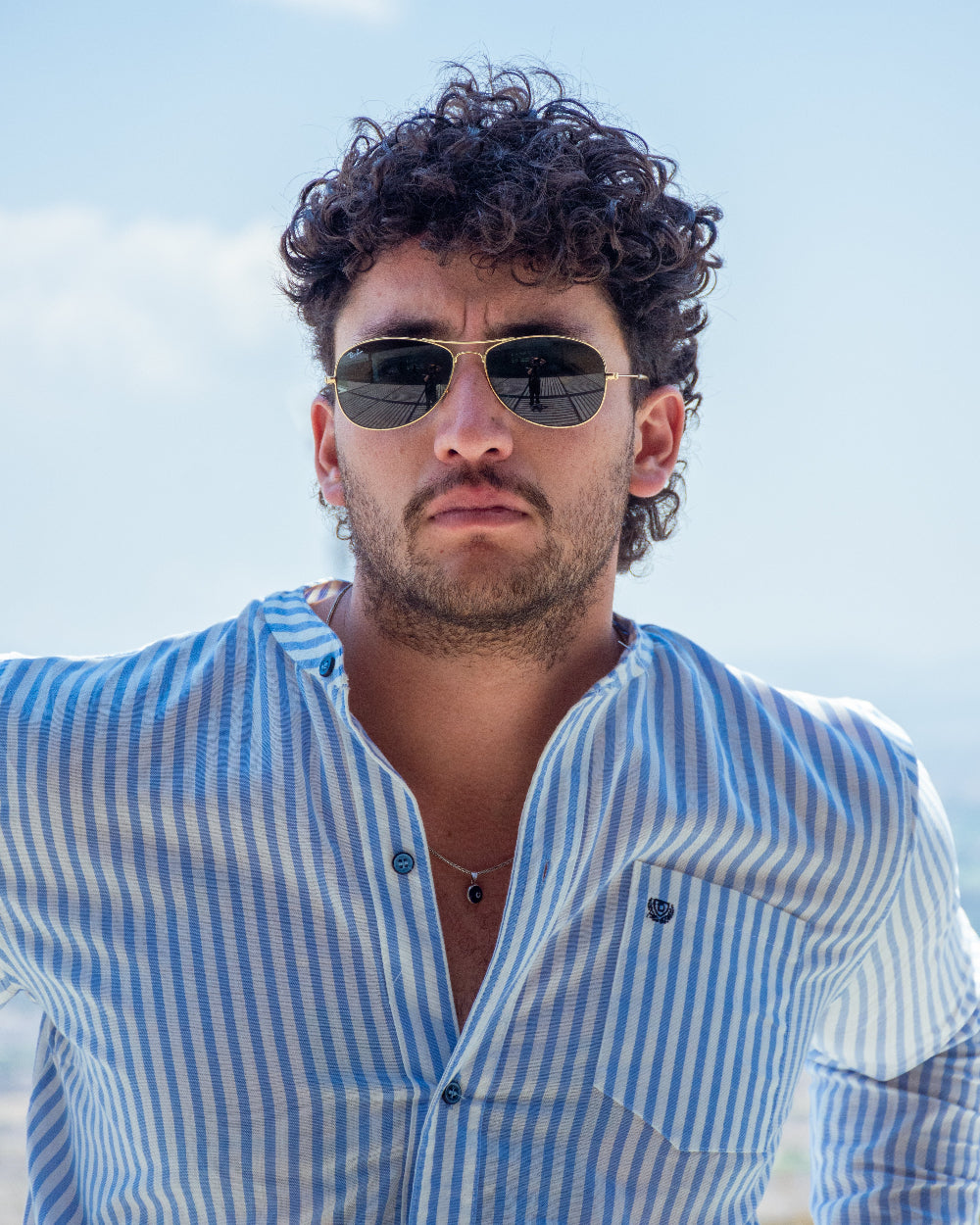 Primer plano de modelo masculino con gafas de sol luciendo camisa de cuello mao a rayas blancas y celestes de la marca Dubrovnik