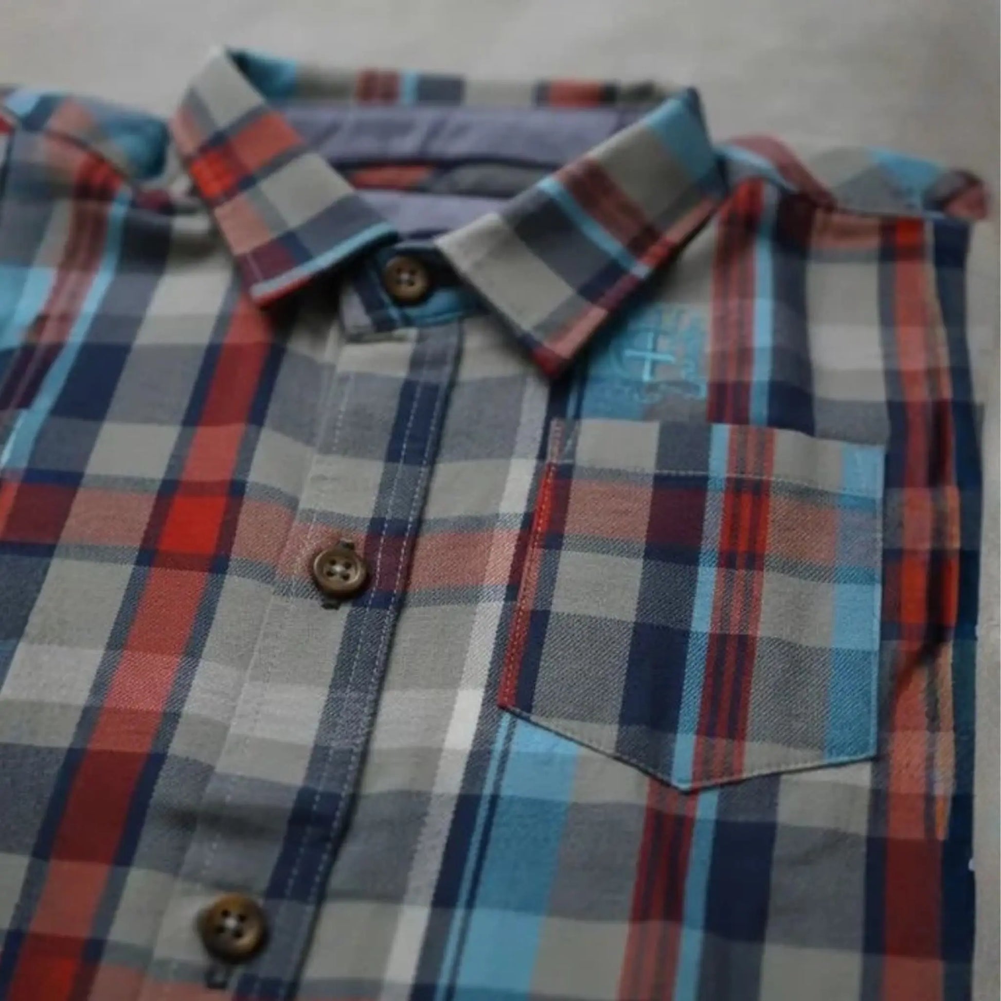 Detalle de costuras y bolsillo de camisa infantil a cuadros en tonos grises, rojos y azules con botones en contraste