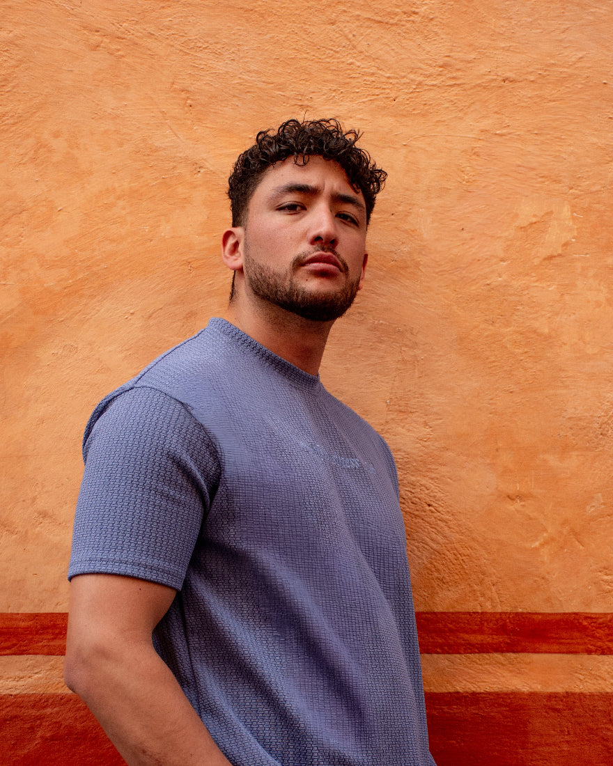 Primer plano de hombre con camiseta azul de tejido nido de abeja y gráfico sutil, posando sobre un fondo naranja cálido con actitud urbana