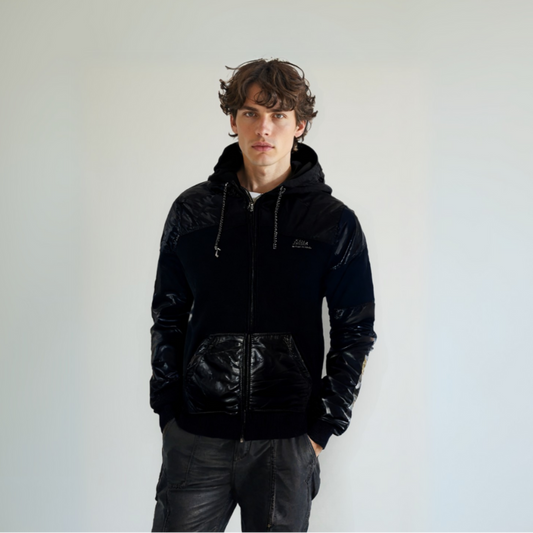 Modelo joven con sudadera negra de cremallera frontal, resaltando el diseño híbrido con paneles de material brillante en hombros, mangas y bolsillos, de la colección exclusiva de Dubrovnik